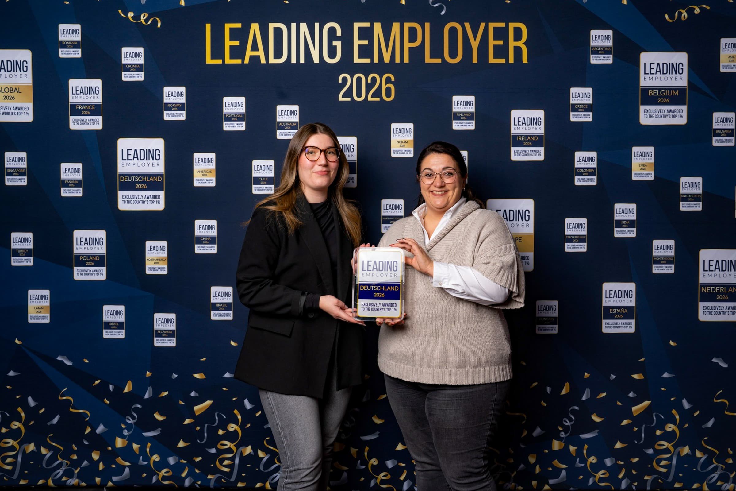 Mandy und Emily aus dem Personalservice der WERTGARANTIE Group stehen vor einer Fotowand und halten gemeinsam den Leading Employer Award in den Händen.