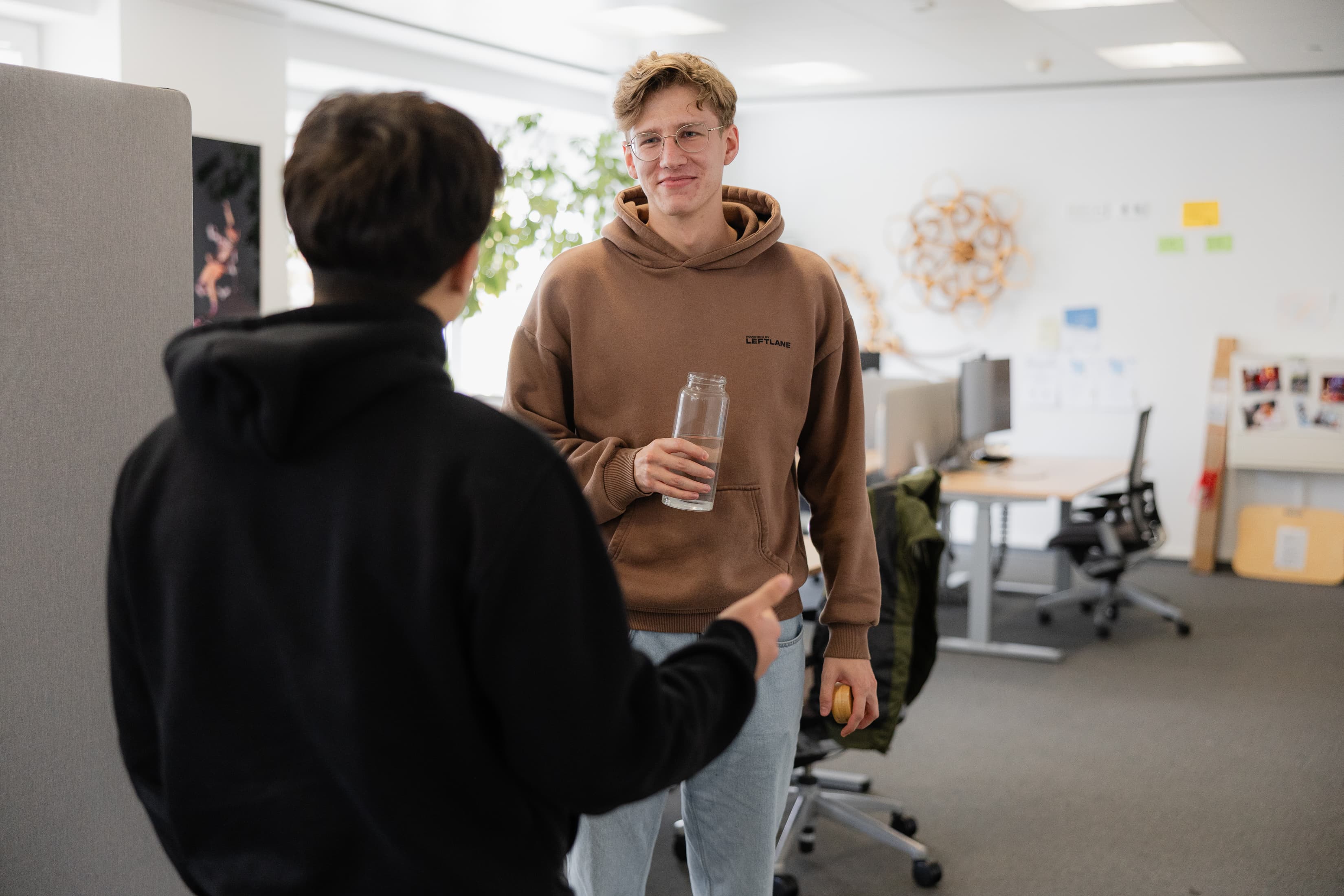 Wertschätzendes Miteinander Unser Dualer Student steht im Büro, schaut in die Kamera und unterhält sich mit einem Kollegen der mit dem Rücken zur Kamera steht.